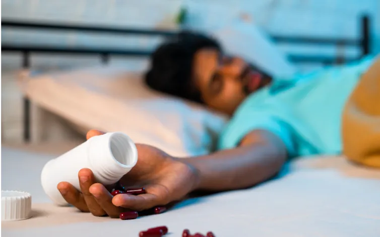 Man lying on back in bed, hand outstretched holding empty pill bottle, pills strewn on floor