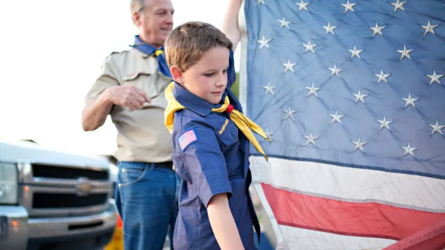 Boy wearing Boy Scout uniform holds American flag under view of adult troop leader
