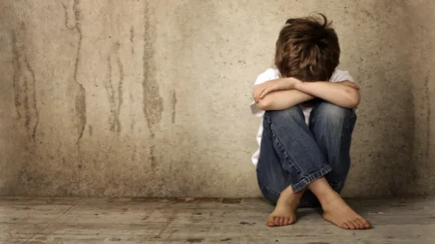 Boy sitting on dirty floor in front of concrete wall with head in his lap